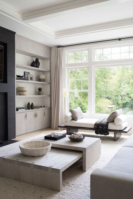 a light-filled greige living room with built-in storag eunits and shelves, a bench, a dup of coffee tables and a greige sofa plus a black fireplace