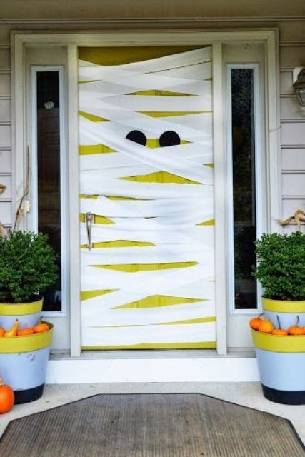a neon neon yellow door styled as a mummy, with pumpkins in planters around is a lovely and playful solution for Halloween