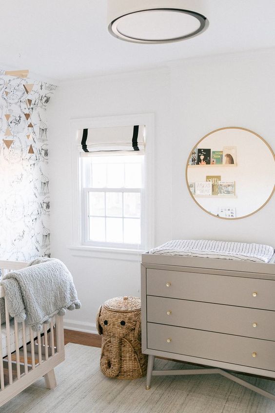 a neutral and pretty nursery with a wallpaper accent wall, greige furniture, a round mirror, ledges with books and an elephant basket for storage
