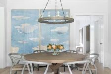 a neutral farmhouse dining room with light-stained wooden beams, a greige round table and chairs, a cool chandelier and a blue artwork on the wall