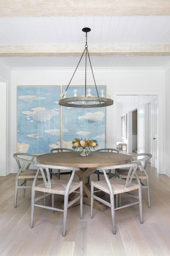 A neutral farmhouse dining room with light stained wooden beams, a greige round table and chairs, a cool chandelier and a blue artwork on the wall