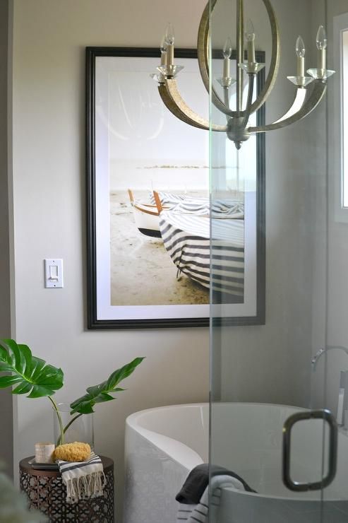 a neutral master bathroom featuring greige walls, a chandelier above a freestanding tub designed and a lovely coastal artwork