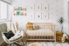 a neutral nursery with a wallpaper accent wall, a light stained crib, jute poufs, a white rocker, a grid gallery wall and open shelves