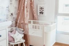 a pretty Scandi nursery with white furniture, a pink canopy with lights, some neutral and pastel layered rugs, a wallpaper accent wall and some artworks
