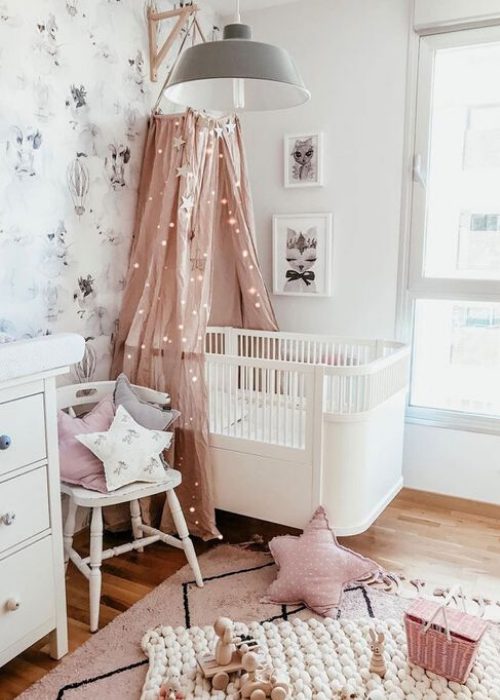a pretty Scandi nursery with white furniture, a pink canopy with lights, some neutral and pastel layered rugs, a wallpaper accent wall and some artworks