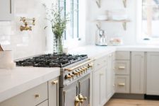 a pretty and refined greige kitchen with shaker cabinets, gold handles and white stone countertops, a white tile backsplash and open shelving