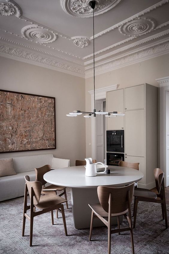 a refined greige dining room with a molded ceiling, a sleek round table, plywood chairs, a bold artwork, a storage unit with built-in appliances