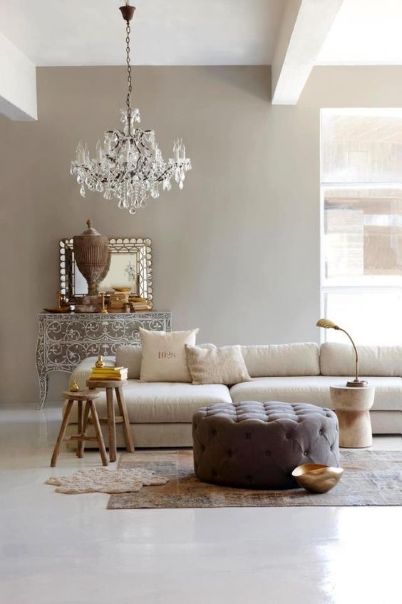 a refined greige living room with a neutral sectional, a taupe tufted pouf, side tables, a crystal chandelier and a beautiful ornated sideboard is wow