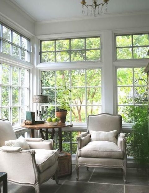 a refined sitting space with large French windows, a round table, neutral vintage chairs, potted greenery and a table lamp is chic