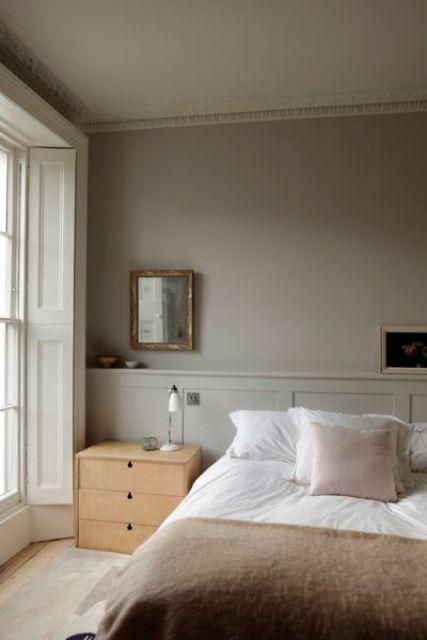 a relaxing greige bedroom with paneling, a bed with neutral bedding, light-stained nightstands, artworks and mirrors plus a mirror with shutters