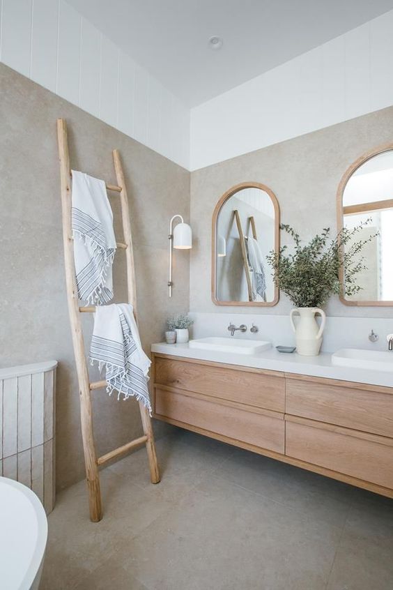 A serene and welcoming greige bathroom with a light stained floating vanity, a ladder, arched windows in wooden frames and a cool sconce
