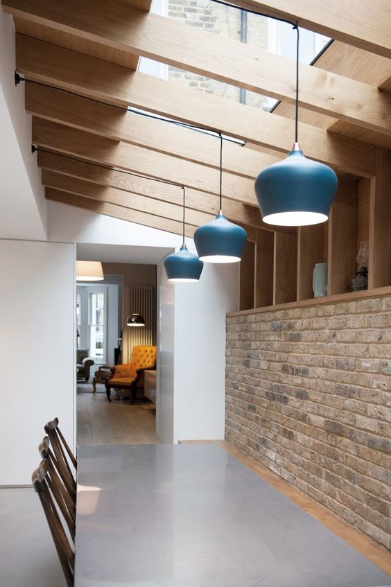 a simple and laconic dining space with a brick wall and open storage shelves, wooden beams and pendant lamps, a dining set lit up with skylights