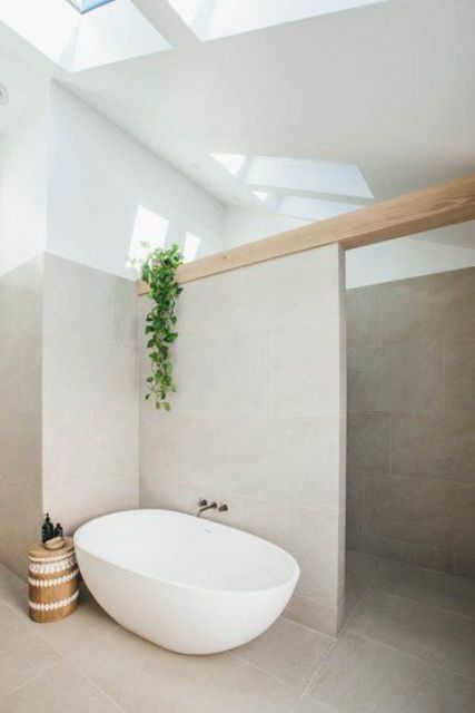 a stylish greige bathroom clad with large scale tiles, a wooden beam, a shower space, an oval tub an a wooden side table plus skylights