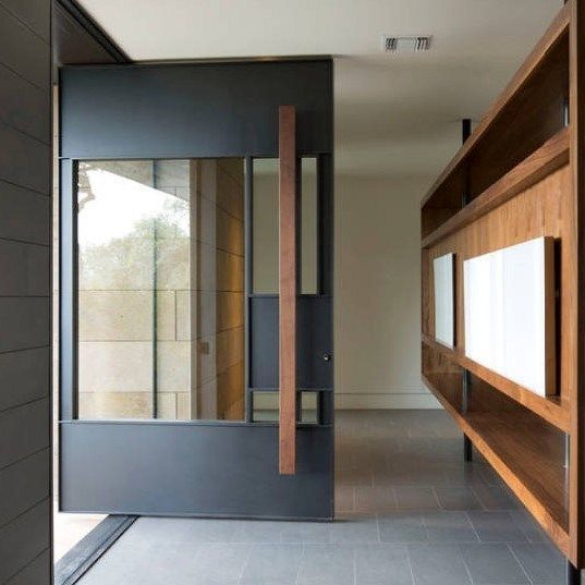 a unique oversized blackened metal front door with a large window right in its center and a wooden handle is gorgeous