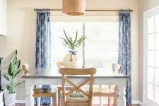 a very simple and relaxed greige dining space with a dining table and stained chairs, blue printed curtains, a pendant lamp