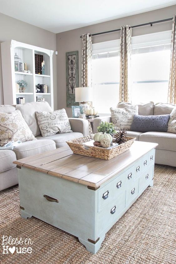 a vintage farmhouse greige living room with neutral sofas, a blue chest coffee table, an open storage bookcase, printed textiles