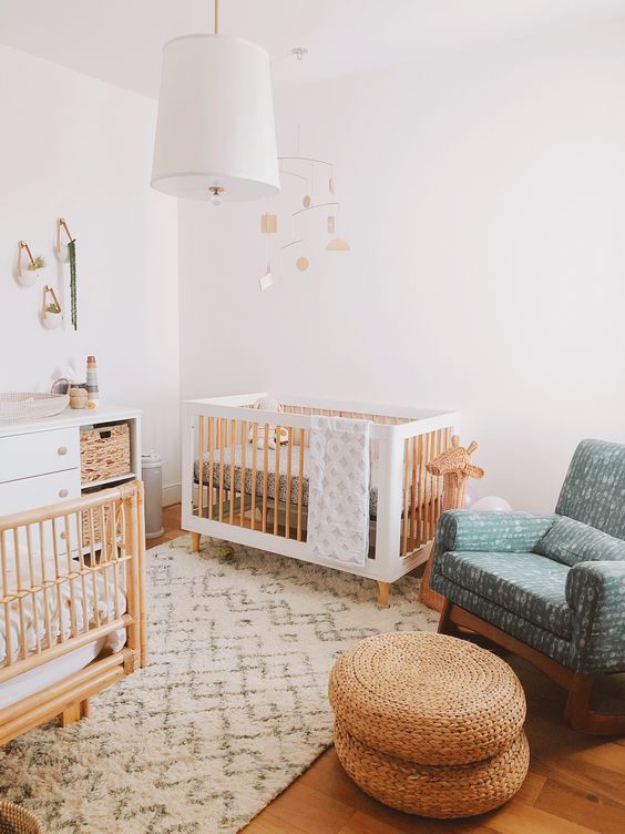 a welcoming Scandi nursery with white and stained furniture, a blue rocker chair, jute poufs, a white dresser and a pendant lamp