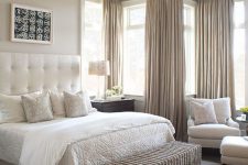 a welcoming greige bedroom with a creamy bed with a statement upholstered headboard, creamy seating furniture, a woven chest bench, greige curtains and a pendant lamp