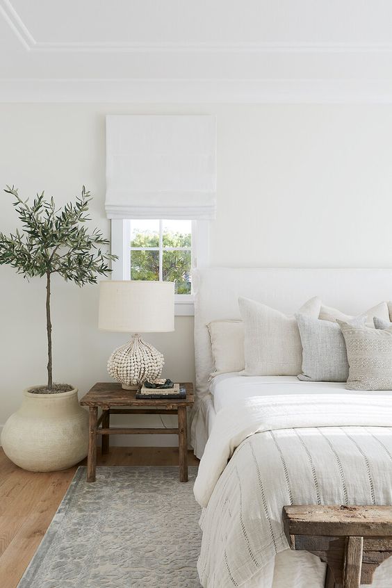 A beautiful all neutral bedroom with a white upholstered bed, a wooden nightstand and a shabby chic bench, a potted plant and all neutral textiles