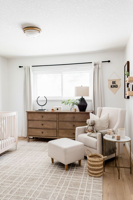 A mid century modern neutral nursery with a white crib, a neutral chair with a footrest, a stained dresser and neutral textiles is welcoming