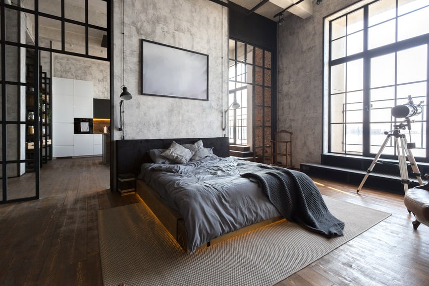 a chic industrial bedroom with concrete walls and a dark stained floor, a black floating bed with lights and grey bedding