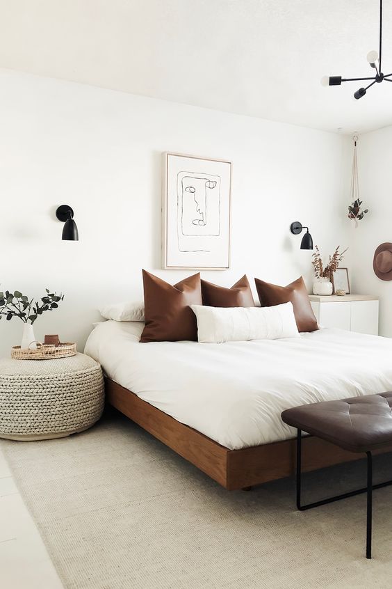 A chic mid century modern bedroom in neutrals, with a rich stained bed with neutral bedding, a leather bench, a woven ottoman and black sconces