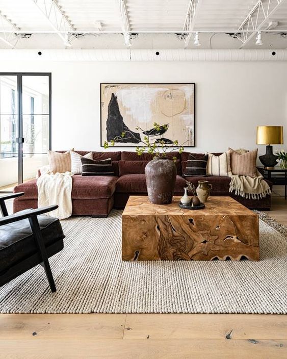 A refined living room with a burgundy sectional, a black chair, a light stained wooden floor, a neutral rug and a rich stained wooden table