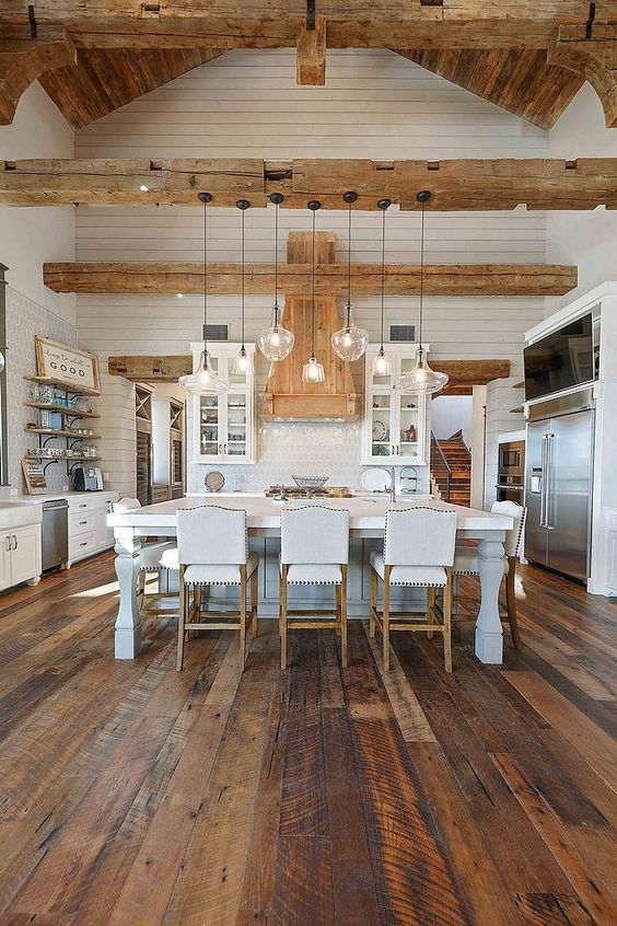 A white farmhouse kitchen with white cabinets, a refined kitchen island, a reclaimed wooden floor, light stained wooden beams and a ceiling