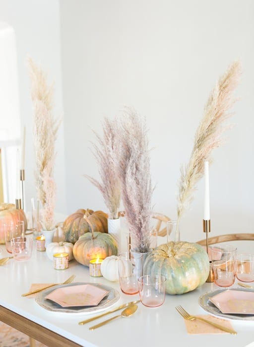 A modern glam Thanksgiving tablescape with pink plates, napkins, glasses, natural and pink grass, heirloom pumpkins and gold cutlery