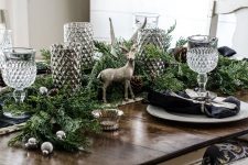 33 a forest glam Christmas tablescape with a greenery runner, silver ornaments, pinecones, glam and shiny candleholders, elegant glasses, dark napkins and silver chargers