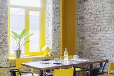 a catchy industrial dining room with brick walls, a dining table, black and yellow chairs, yellow window frames and a radiator on the wall