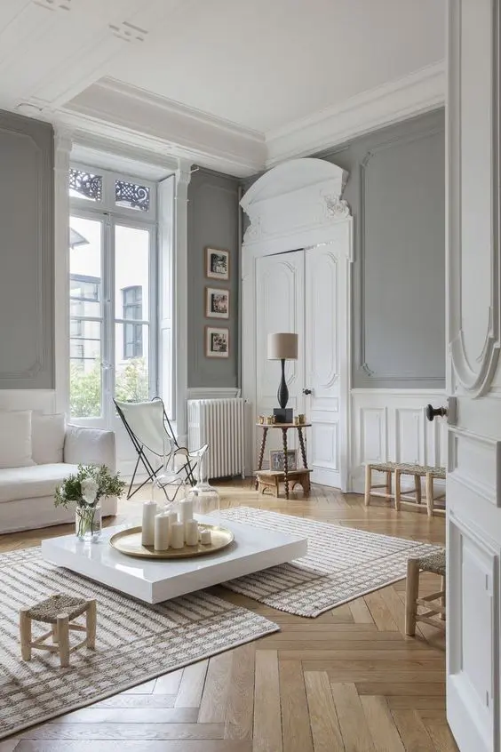 A jaw dropping neutral Parisian space with grey walls, white wainscoting, crown molding, white furniture and neutral colored chairs and stools