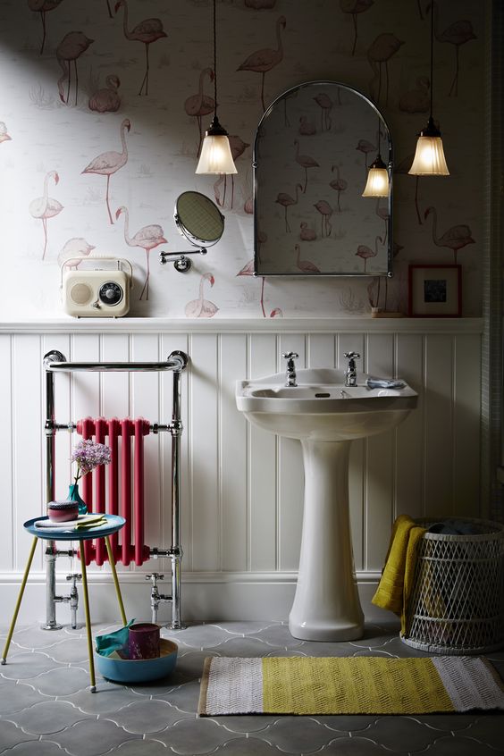 A lovely bathroom with flamingo printed wallpaper and white wainscoting, a free standing sink, a small pink radiator, a bold rug and pendant lamps