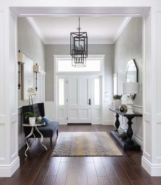 a sophisticated entryway with greige walls, white wainscoting and the door, a black upholstered bench, a black console table and some artworks and a mirror