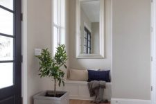 a stylish greige entryway with a built-in paneled bench, a planter with a tree and a tan and navy pillow is an elegant space