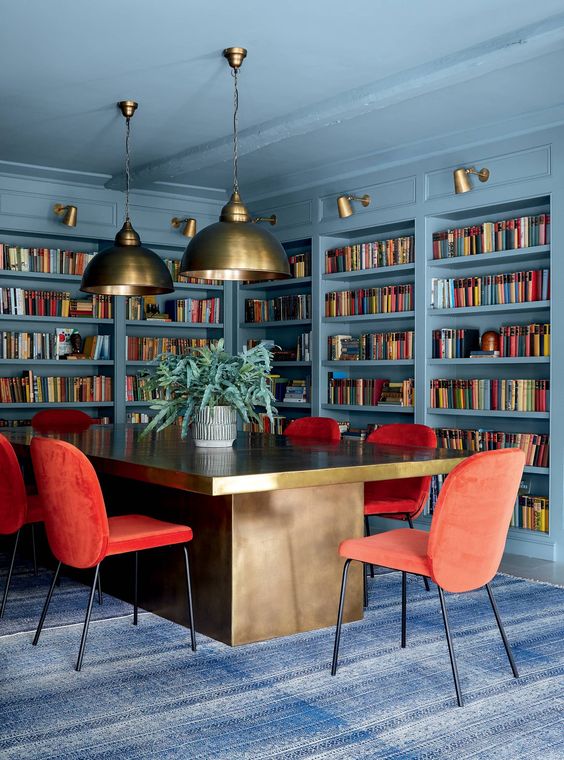 A beautiful stone blue home library and home office with lots of built in shelves, a large refined desk, red chairs and pendant lamps