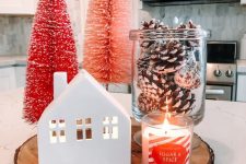 23 a small Christmas arrangement of a mini house, a jar with snowy pinecones, a candle, pink and red bottle brush Christmas trees