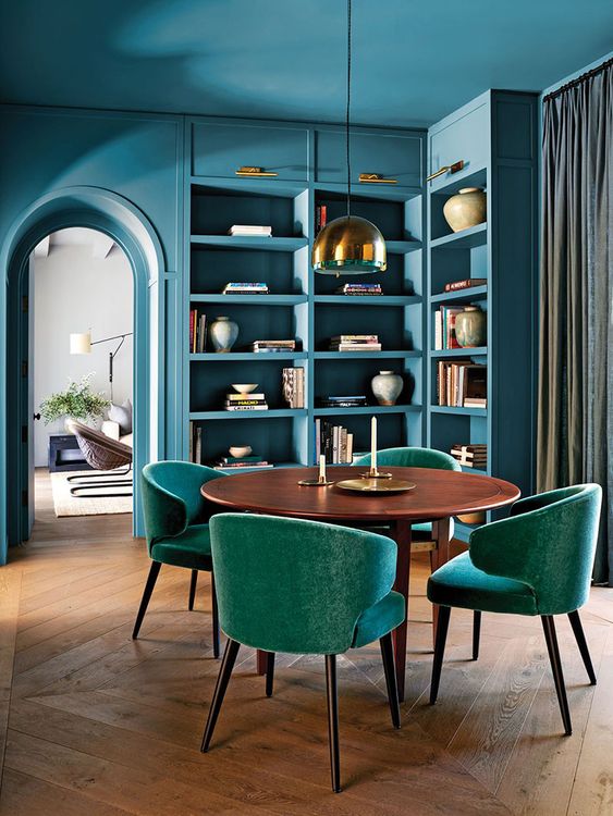 A fabulous stone blue dining space with built in shelves and storage units, a stained dining table, teal chairs and a pendant lamp