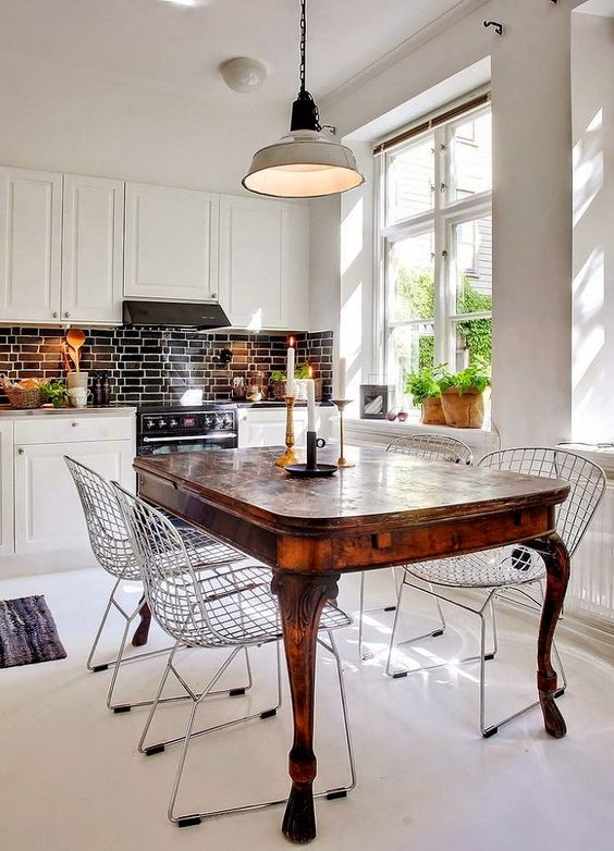 A white eat in kitchen with a black tile backsplash, a rich stained dining table and modern wire chairs for a contrast