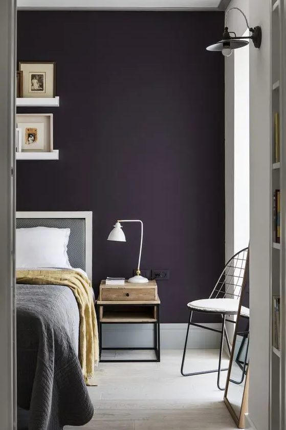a stylish moody bedroom with an aubergine accent wall, grey and white furniture, a small nightstand with a table lamp and ledges with artworks