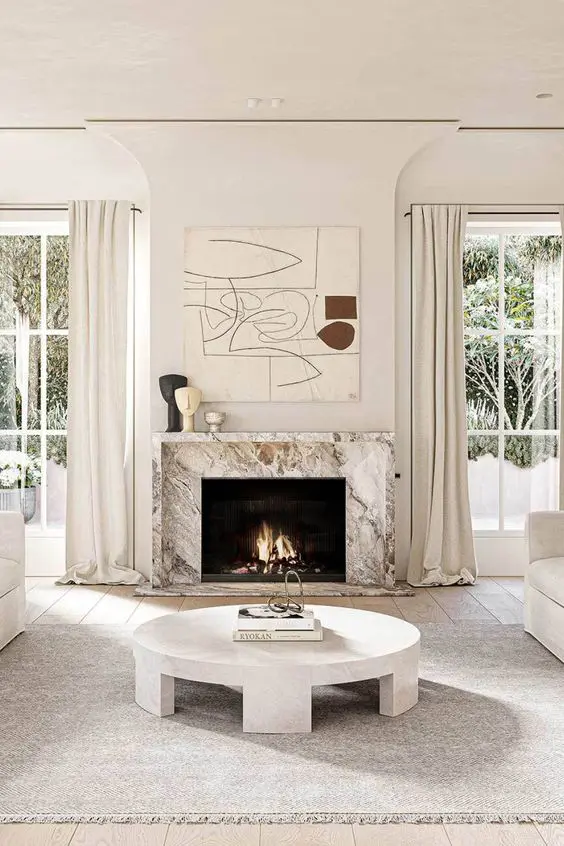 a beautiful and airy living room done in cremay shades, with a white marble fireplace, a statement artwork and a round coffee table