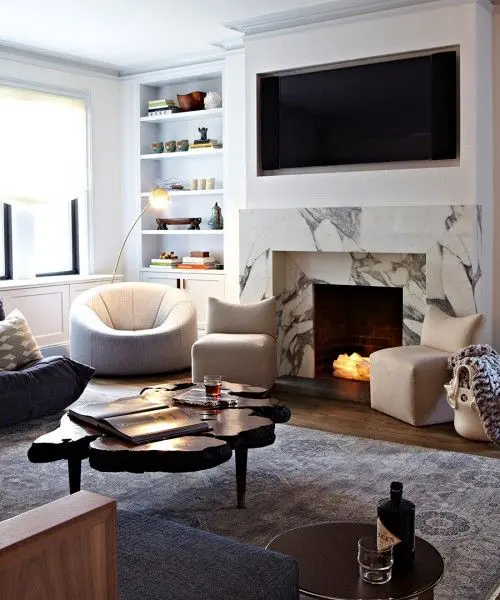 A beautiful and catchy modern living room with a built in white marble fireplace, a built in TV, built in shelves, white and grey furniture and a wood slice table