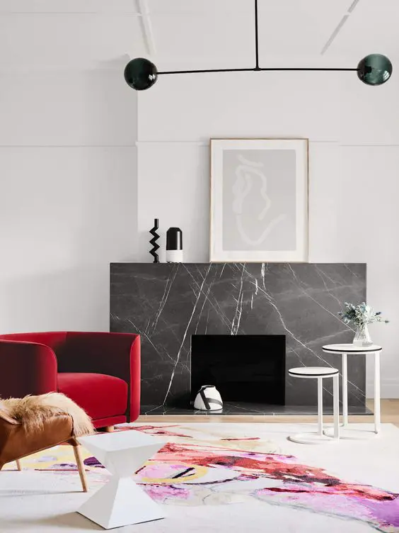 a bold modern space with a fireplace clad with grey marble, a red chair and a leather one, a bold rug, a black chandelier and some art