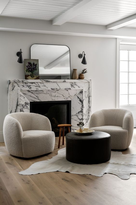 a chic contemporary nook with a white marble fireplace, neutral curved chairs, a black leather pouf, a stool and some art and decor