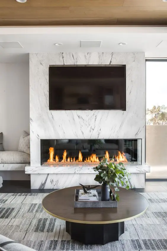a chic modern space with a fireplace clad with white marble and a TV, a built-in bench, a round table and a statement rug