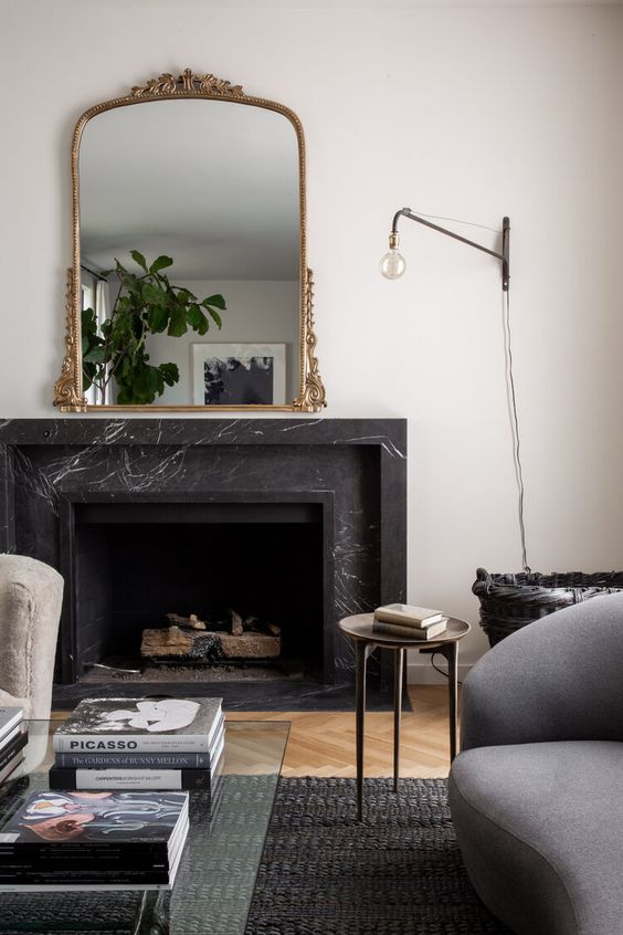 a chic space with a black marble fireplace, neutral and grey seating furniture, a sconce, a mirror with an adorned frame and greenery