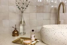 a gorgeous bathroom with a vintage feel, with neutral zellige tiles, a terrazzo sink, a white stone countertop and brass fixtures