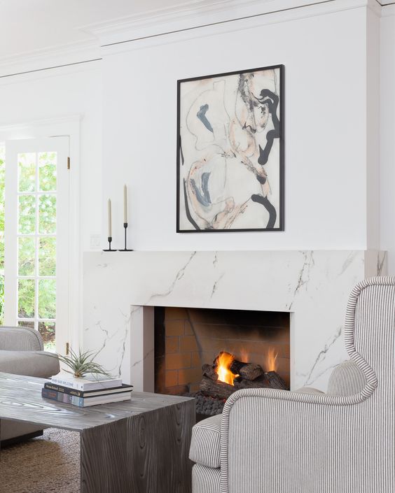 A jaw dropping fireplace nook with a white marble fireplace, striped chairs, a wooden coffee table and a bold artwork