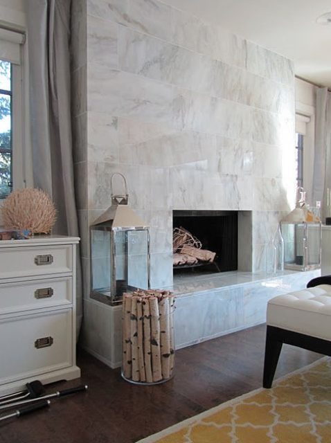 A large non working fireplace clad with white marble tiles all over, with candle lanterns and firewood in a vase