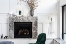 a sophisticated nook with a marble clad fireplace, a grey ottoman, a green tufted chair, some decor on the mantel and a floor lamp